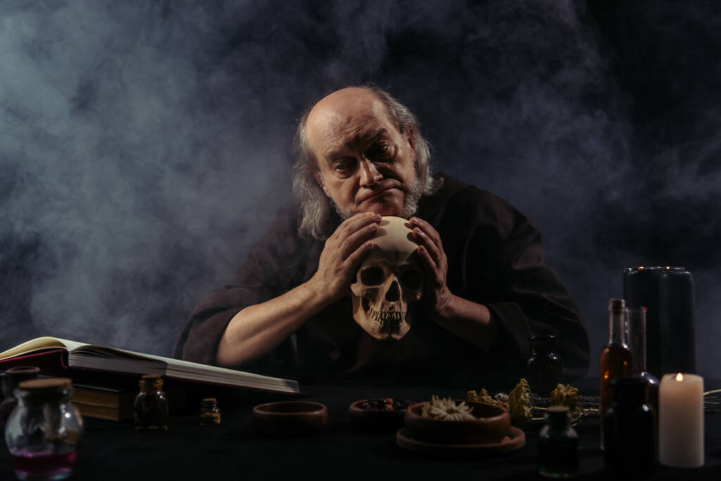 odrazený alchymista držící lebku v blízkosti složek a magické kuchařky na černém pozadí s kouřem - Fotografie, Obrázek