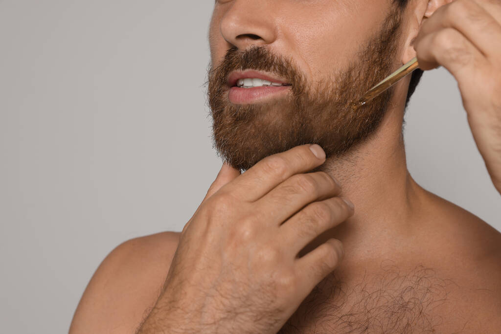 Man applying oil onto beard on grey background, closeup - Fotografie, Obrázek