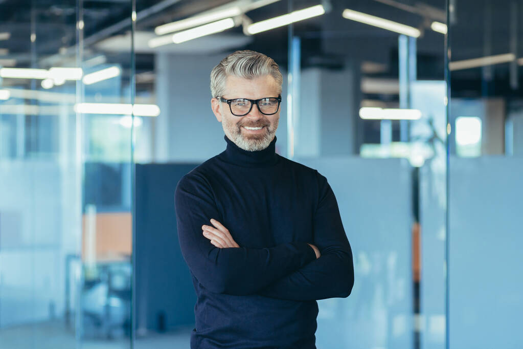 Portrait of successful businessman in modern office with crossed arms, gray haired male investor smiling and looking at camera, boss in glasses and jeans - 写真・画像