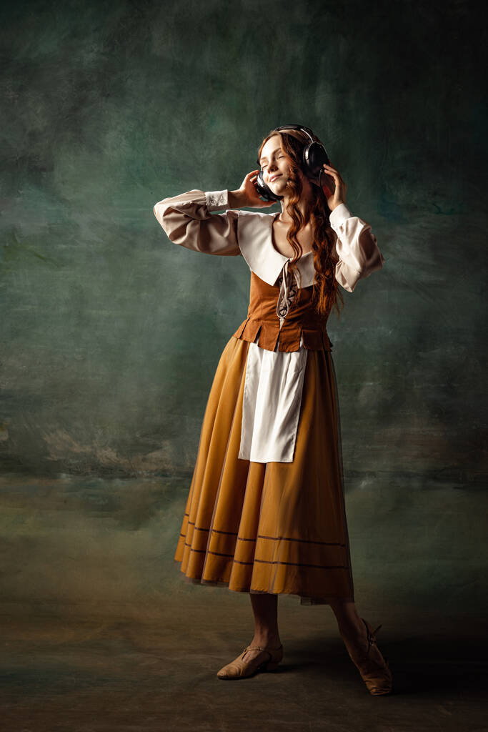 Music. Portrait of charming redhair girl with long curly hair like girl of renaissance eras, lady-peasant isolated on dark background. Comparison of eras, beauty, history, art, creativity. - Foto, imagen