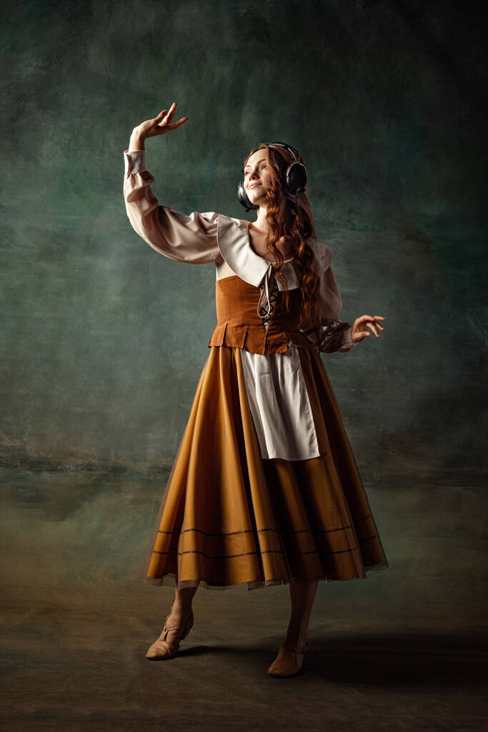 Dancing. Young charming redhair girl with long curly hair like girl of renaissance eras isolated on dark background. Comparison of eras, beauty, history, art, creativity. - Foto, Imagem