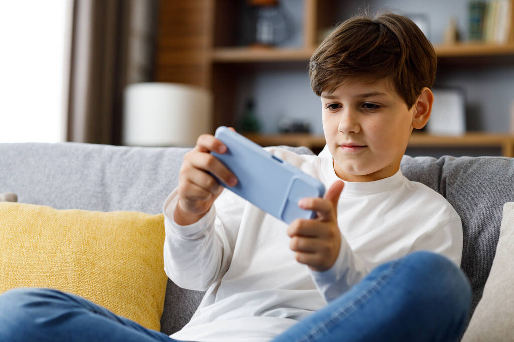 Cheerful young boy playing video game on smartphone sitting on sofa at home. Cute teenager playing in car racing game on mobile phone. Spending time at home, leisure, relax, happy carefree childhood. - Fotografie, Obrázek