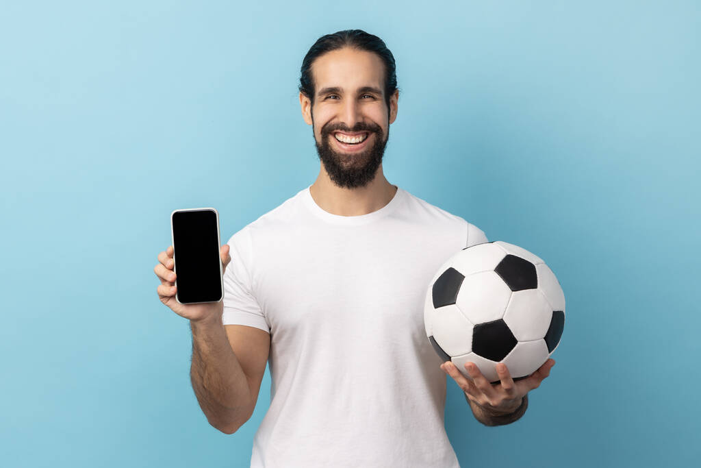 Portrait of happy man with beard wearing white T-shirt holding soccer ball and smartphone empty black display, ticket booking for championship. Indoor studio shot isolated on blue background. - Fotografie, Obrázek