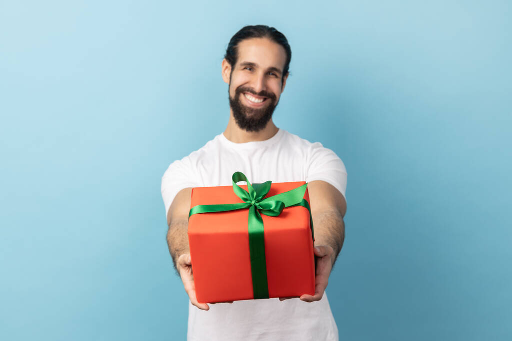 Portrait of handsome delighted man with beard wearing white T-shirt giving wrapped gift box and smiling at camera, happy holiday. Indoor studio shot isolated on blue background. - Foto, imagen