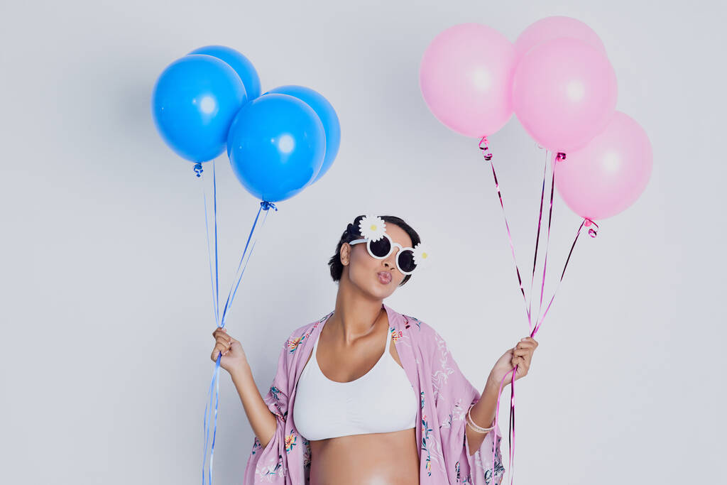 Will it be a girl or a boy. Studio shot of a beautiful young pregnant woman holding blue and pink balloons against a gray background - Foto, afbeelding