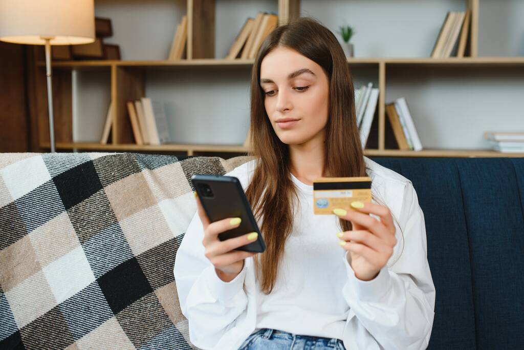 Щасливий підліток робить покупки онлайн, сидячи на дивані вдома. Молода жінка вставляє номер кредитної картки на веб-сайт зі своїм смартфоном
 - Фото, зображення