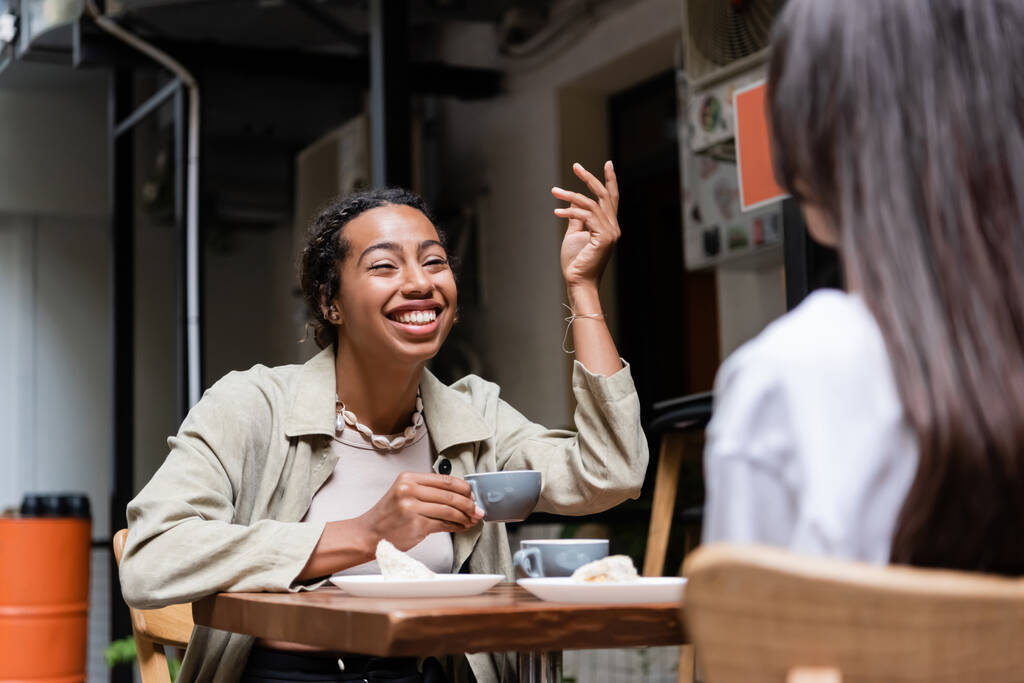 Αφρο-αμερικανίδα γυναίκα κρατώντας φλιτζάνι καφέ και μιλώντας με θολή φίλη στη βεράντα του καφέ  - Φωτογραφία, εικόνα