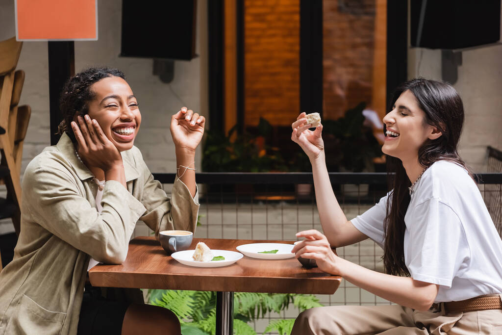 Смеющиеся многонациональные девушки разговаривают возле кофе и десертов на террасе кафе  - Фото, изображение
