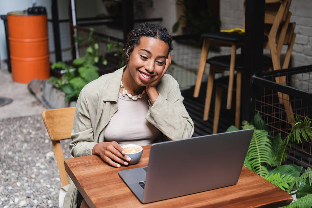 Smiling african american woman holding cup of coffee and looking at laptop in outdoor cafe  - Foto, afbeelding