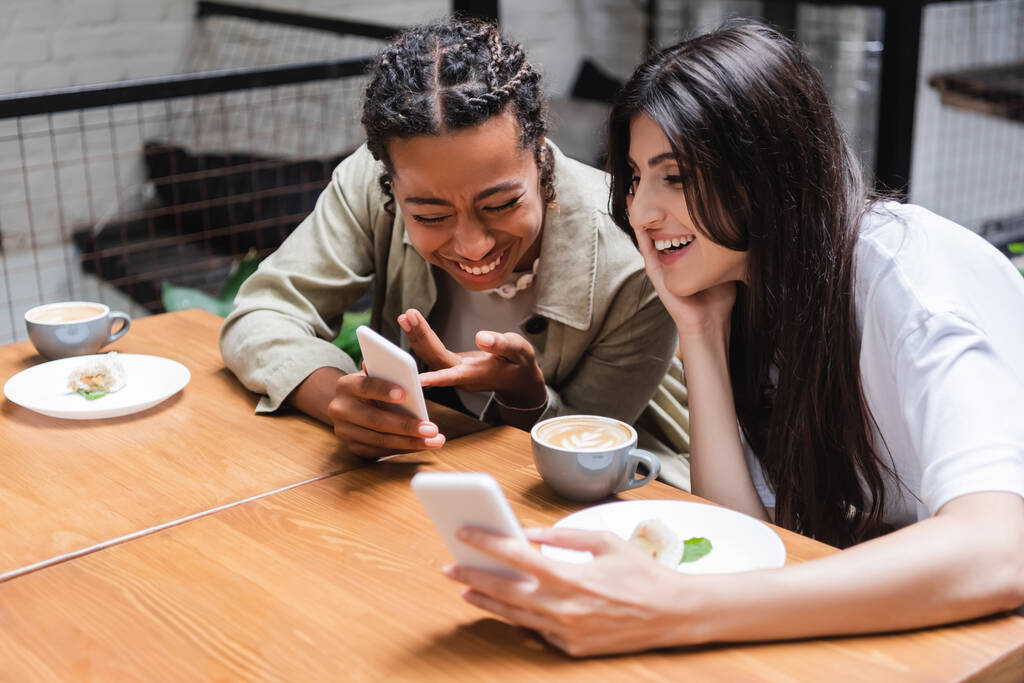 Positive multiethnic girlfriends using smartphones near coffee and desserts in outdoor cafe  - Zdjęcie, obraz