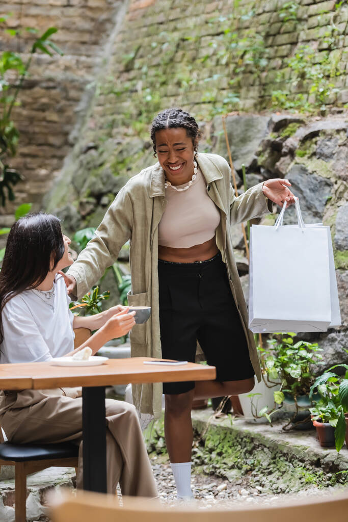 Positive african american woman holding shopping bags near friend with coffee in outdoor cafe - Photo, Image