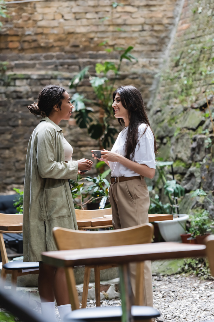Side view of multiethnic girlfriends holding coffee cups while talking on terrace of cafe  - Photo, Image