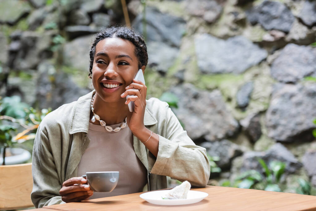 Smiling african american woman talking on smartphone near coffee and dessert on terrace of cafe  - Foto, afbeelding