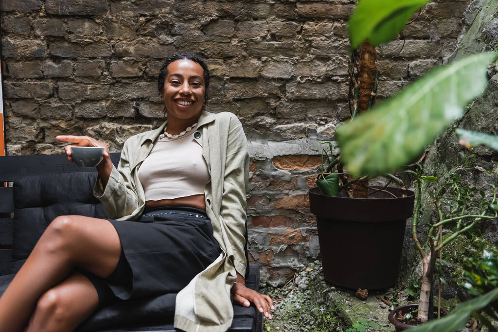 Cheerful african american woman holding cup of coffee near plants on terrace of cafe  - Photo, Image