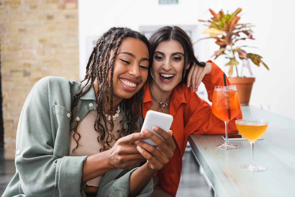 Позитивные многонациональные друзья используют смартфон рядом с коктейлями в кафе  - Фото, изображение