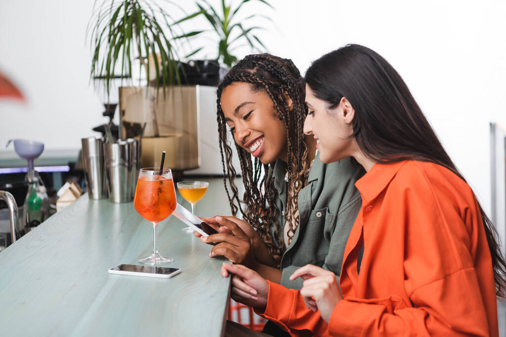 Cheerful multiethnic girlfriends using smartphone near cocktails in cafe  - Photo, image
