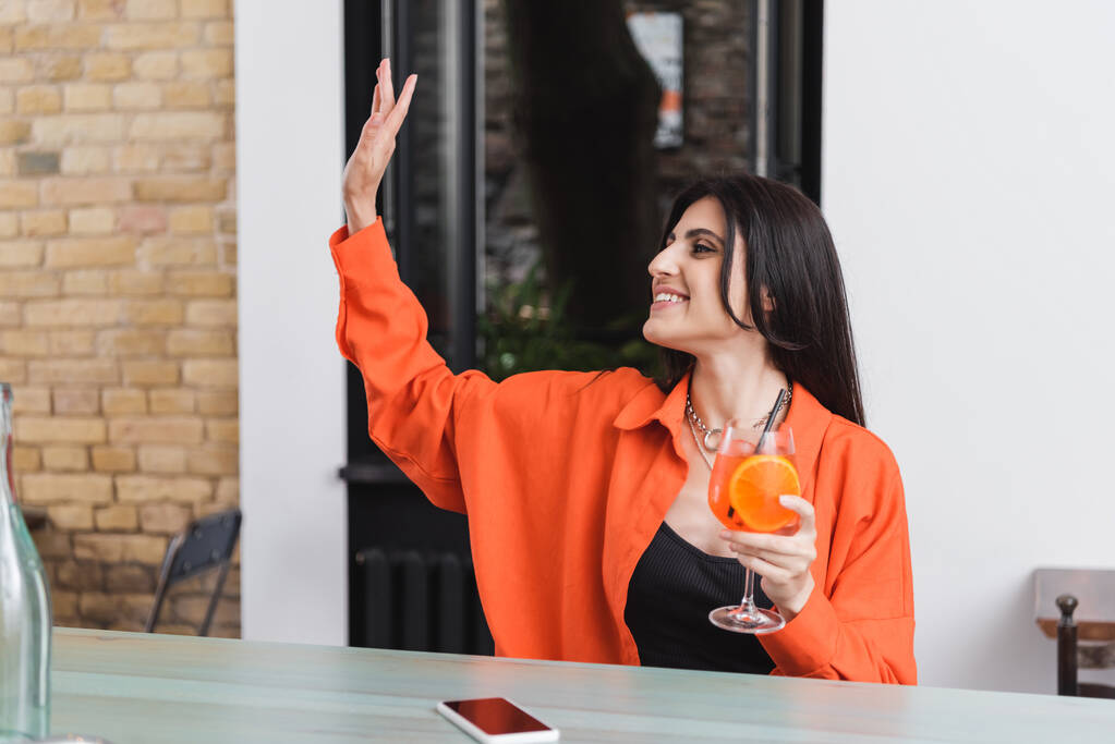 Smiling woman holding cocktail and waving hand near smartphone in cafe  - Fotoğraf, Görsel