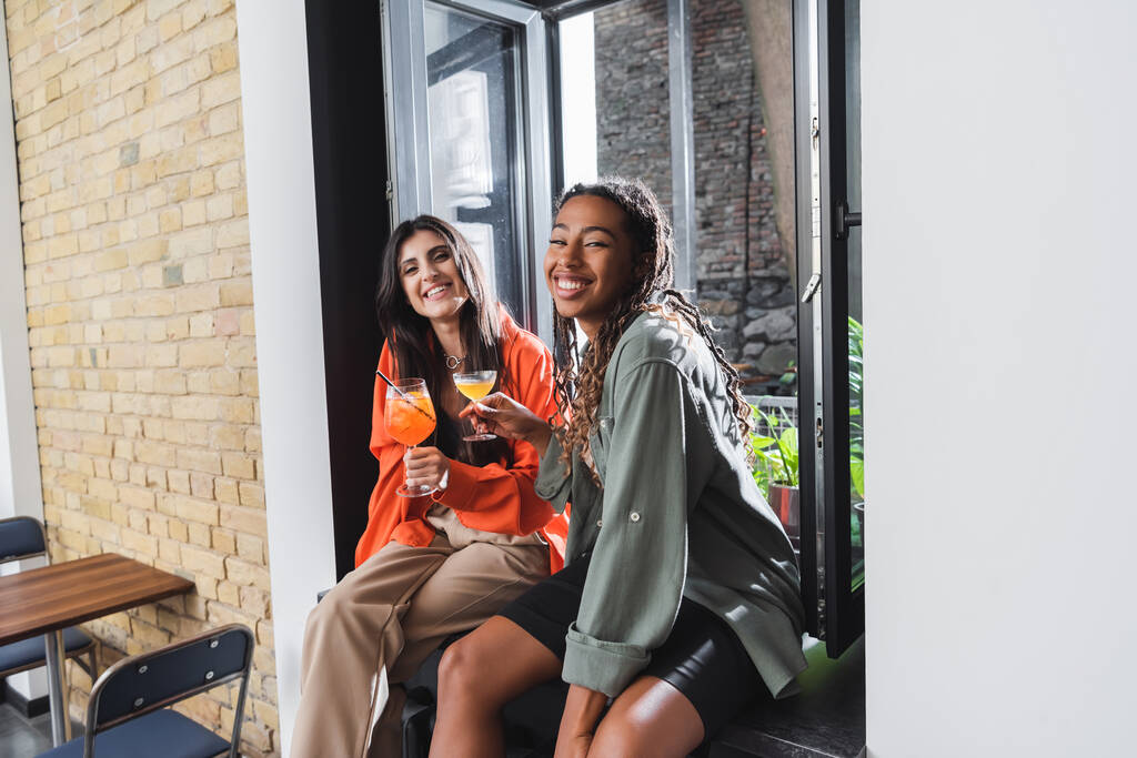 Усміхнені багатоетнічні подружки тримають коктейлі і дивляться на камеру в кафе
  - Фото, зображення
