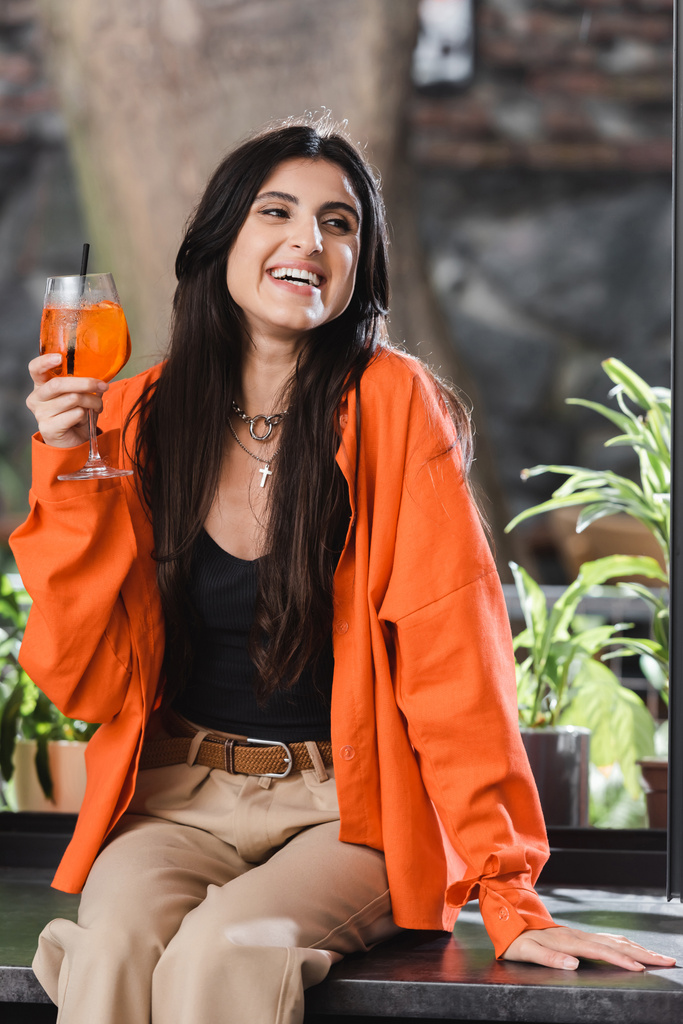 Positive woman holding cocktail and looking away on windowsill in cafe  - Photo, Image