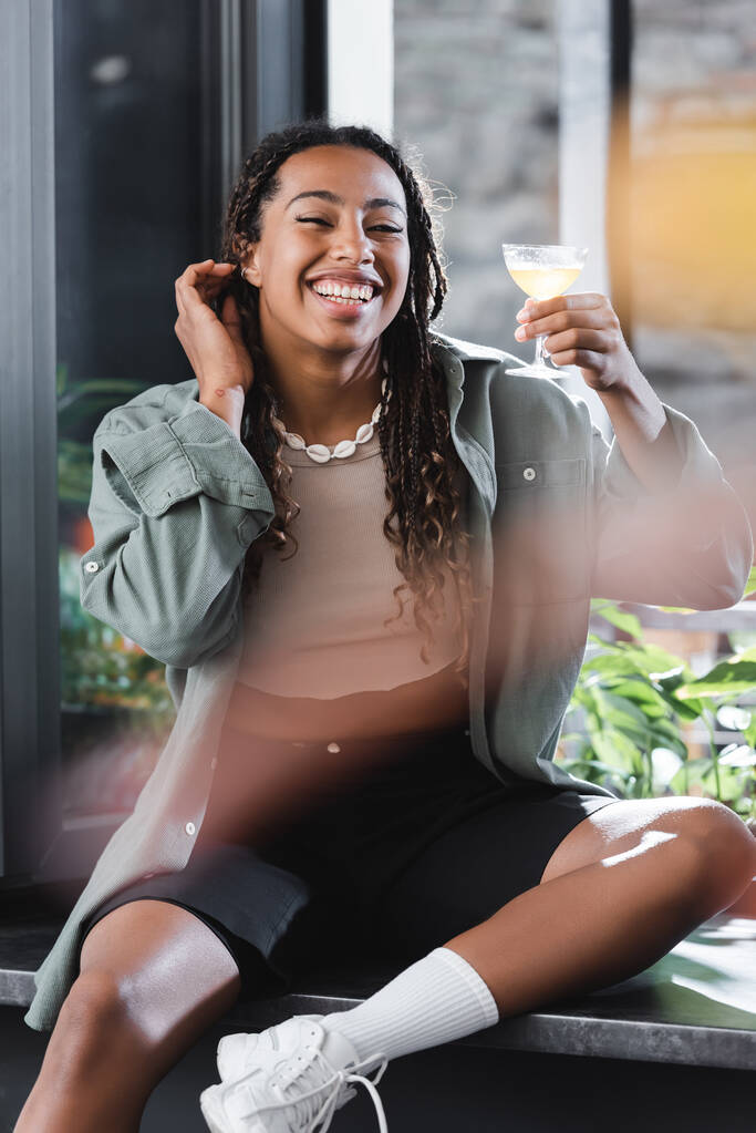 Happy african american woman holding cocktail while sitting near window in cafe  - 写真・画像