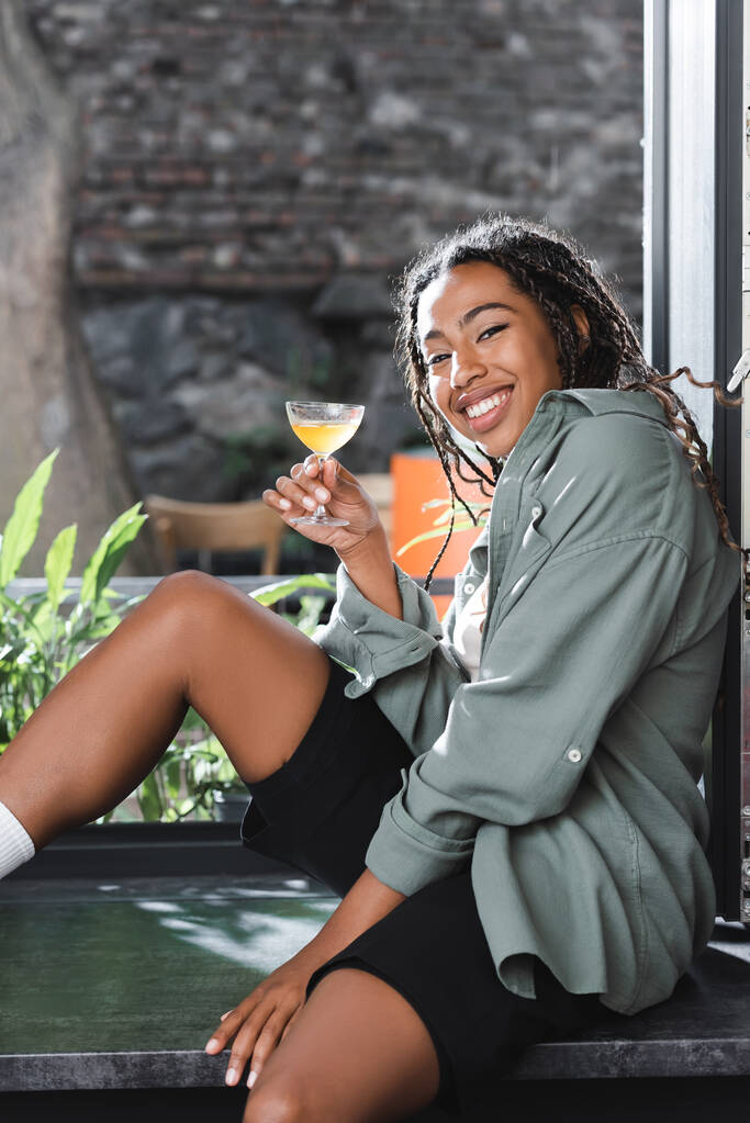 Positive african american woman holding cocktail and looking at camera on windowsill in cafe  - Foto, Imagem