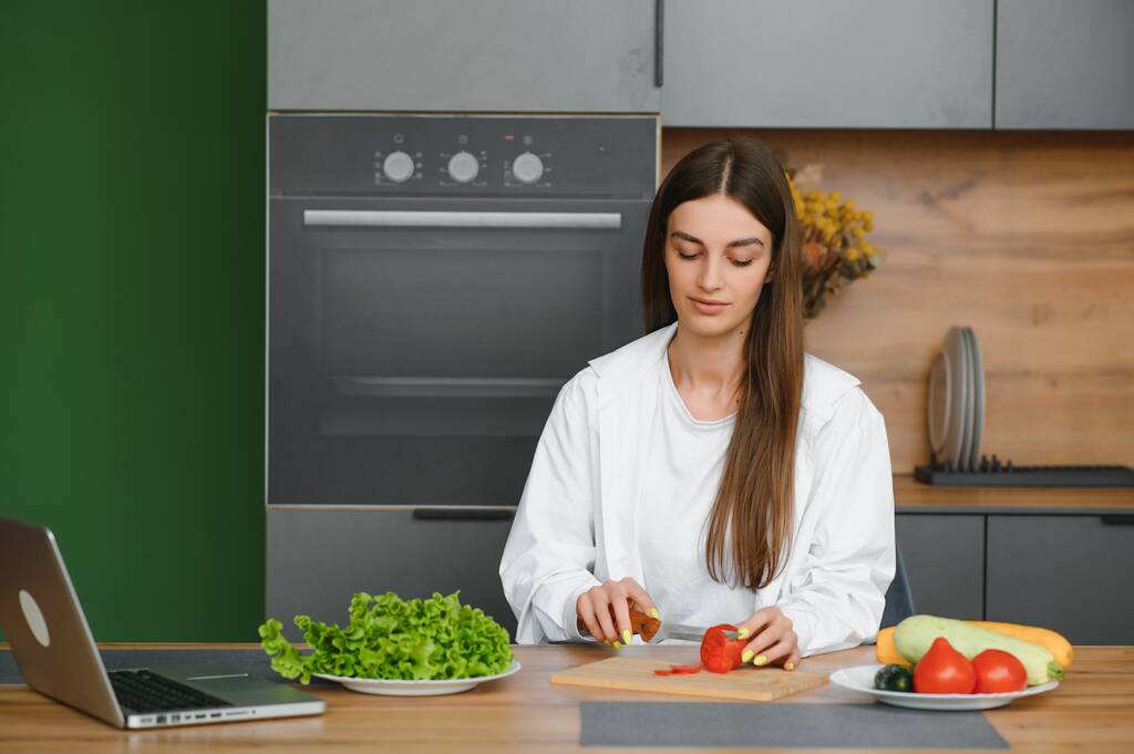Женщина смотрит кулинарные каналы и готовит салат дома. Она режет овощи на разделочной доске и смотрит на экран ноутбука в скандинавском стиле кухни
 - Фото, изображение