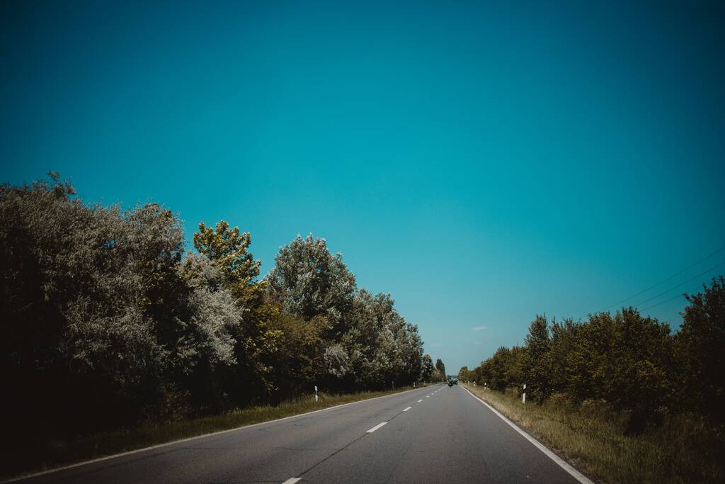 Ένας μακρύς ασφαλτοστρωμένος δρόμος μέσα από ένα αγροτικό χωράφι με δέντρα κάτω από έναν γαλάζιο ουρανό - Φωτογραφία, εικόνα