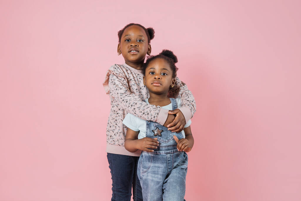 Zwei fröhliche lustige Kinder, lachende kleine afrikanische Mädchen, in pastellfarbener Freizeitkleidung, posieren auf rosa Wandhintergrund. Zwei Kinder Schwestern umarmen einander. - Foto, Bild
