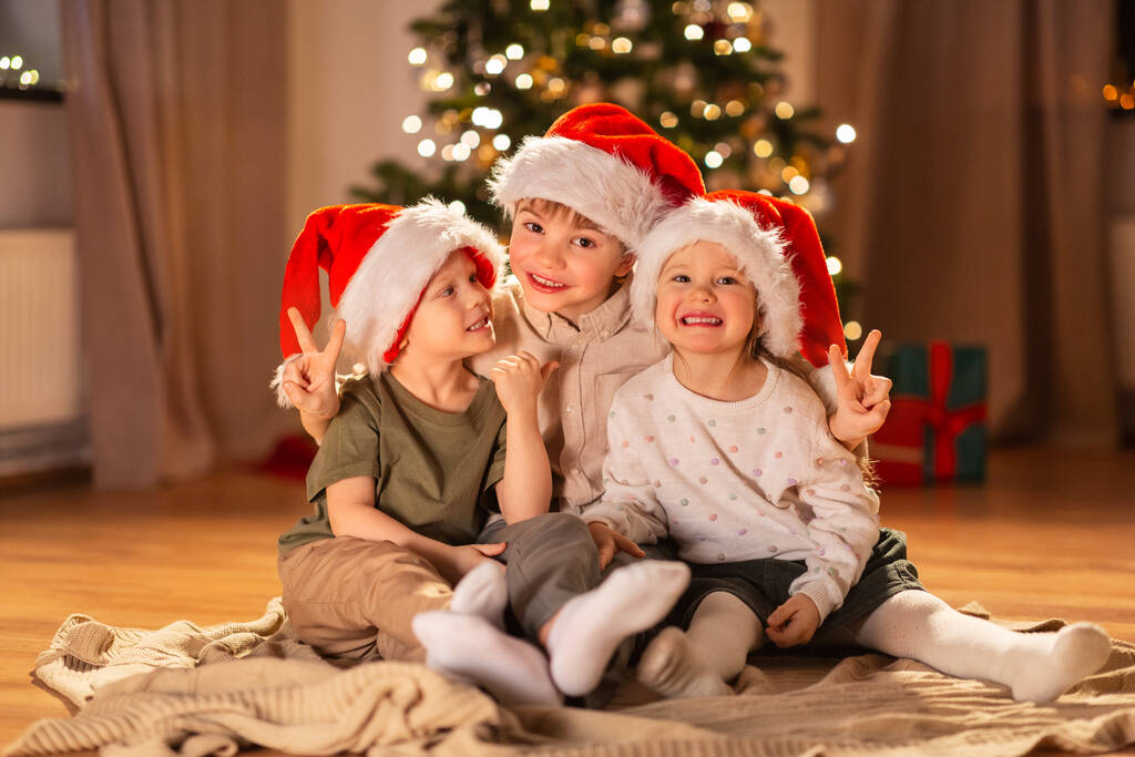 christmas, winter holidays and childhood concept - happy little girl and boys in santa hats showing peace gesture on floor at home - 写真・画像