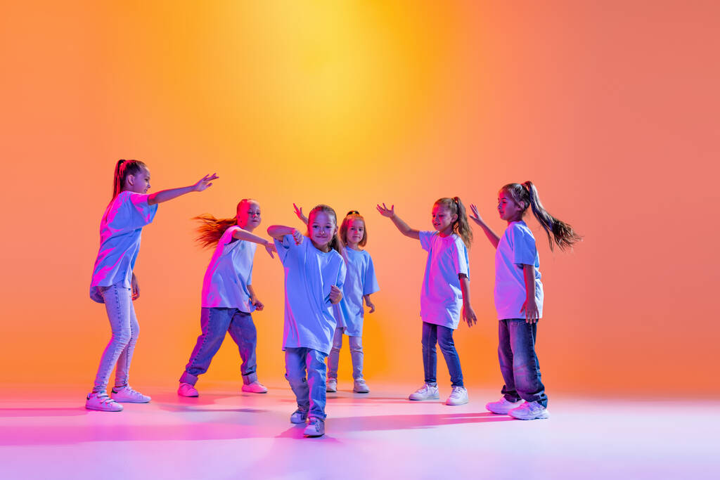 Хип-хоп танец, уличный стиль. Счастливые дети, маленькие активные девочки в повседневной одежде танцы изолированы на оранжевом фоне в фиолетовом неоновом свете. Концепция музыки, моды, искусства - Фото, изображение