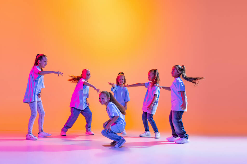 Joy, fun and happiness. Portrait of cheerful, active little girls, happy kids dancing isolated on orange background in neon light. Concept of music, fashion, art, childhood, hobby. - Fotografie, Obrázek