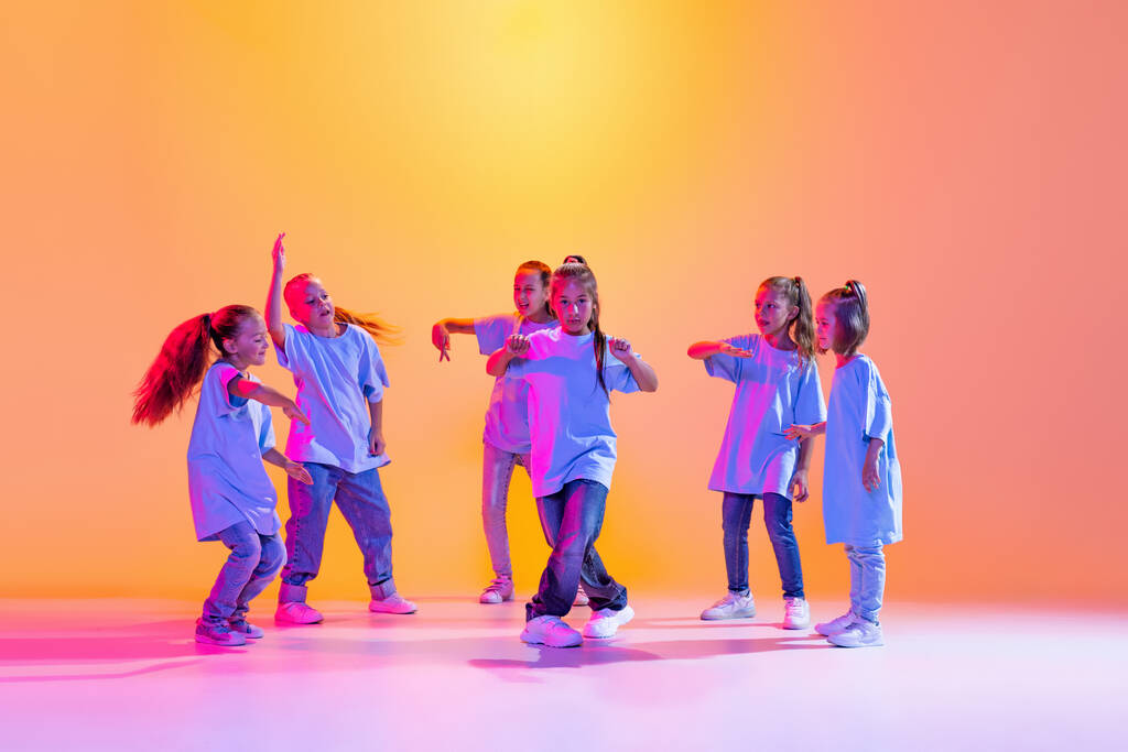Счастливые дети танцуют. Группа детей, маленьких девочек в спортивной повседневной одежде, танцующих в хореографическом классе изолированы на оранжевом фоне в фиолетовом неоновом свете. Концепция музыки, моды - Фото, изображение