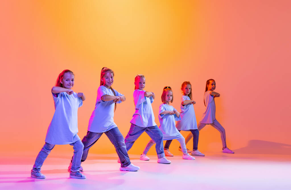 Joy, fun and happiness. Portrait of cheerful, active little girls, happy kids dancing isolated on orange background in neon light. Concept of music, fashion, art, childhood, hobby. - Photo, Image