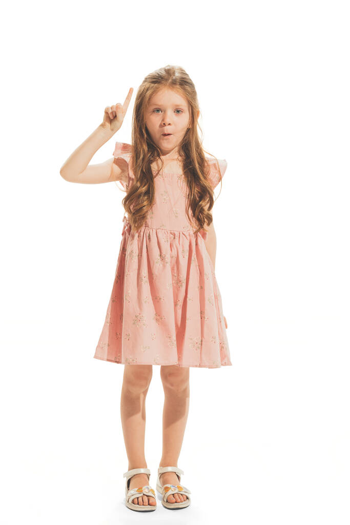 Portrait of beautiful little girl in stylish dress posing, raising finger up isolated on white studio background. Concept of emotions, facial expression, childhood, game, education. Copy space for ad - Foto, afbeelding