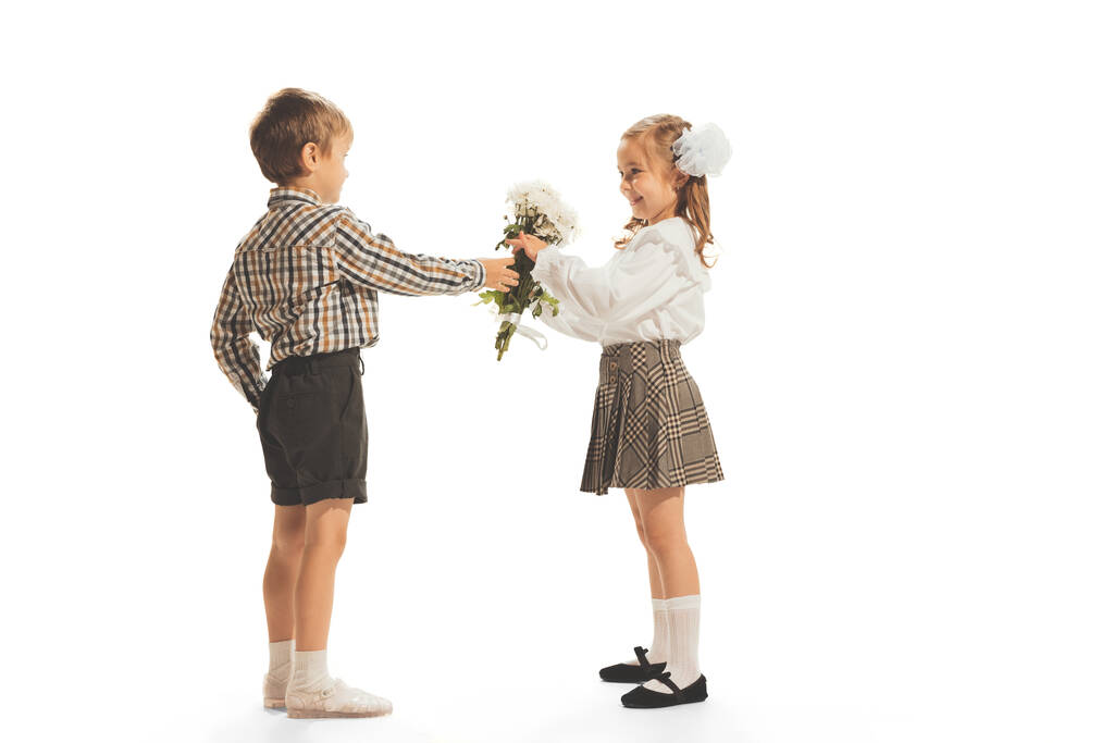 Portrait of children, boy giving girl flowers isolated over white studio background. Retro fashion. Concept of childhood, emotions, style, game, school, fun, education. Copy space for ad - Foto, imagen