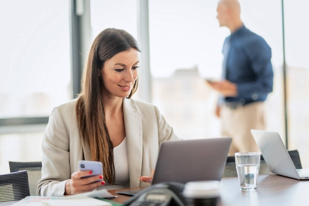 Shot of confident businesswoman using her mobile phone and laptop while sitting at office desk and working. Unrecognizable businessman standing at the background.  - Valokuva, kuva