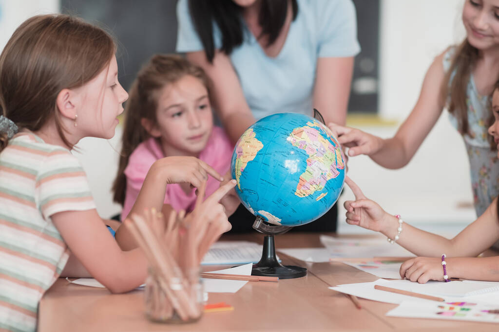 Female teacher with kids in geography class looking at a globe. Side view of a group of diverse happy school kids with a globe in the classroom at school. High quality photo - Fotó, kép