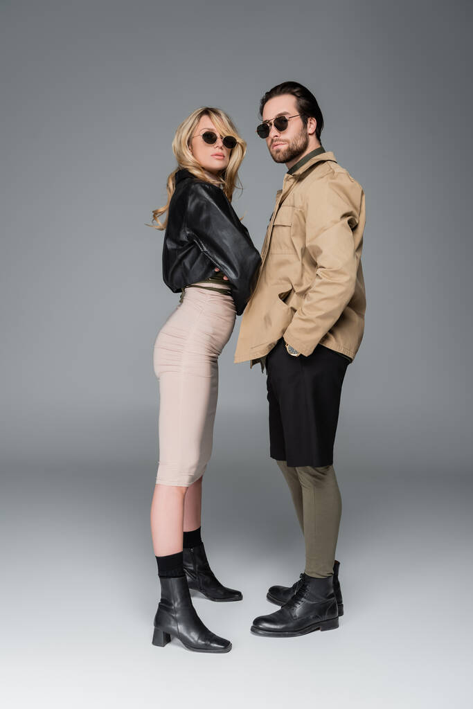 full length of stylish couple in trendy outfits and sunglasses posing together on grey - Zdjęcie, obraz