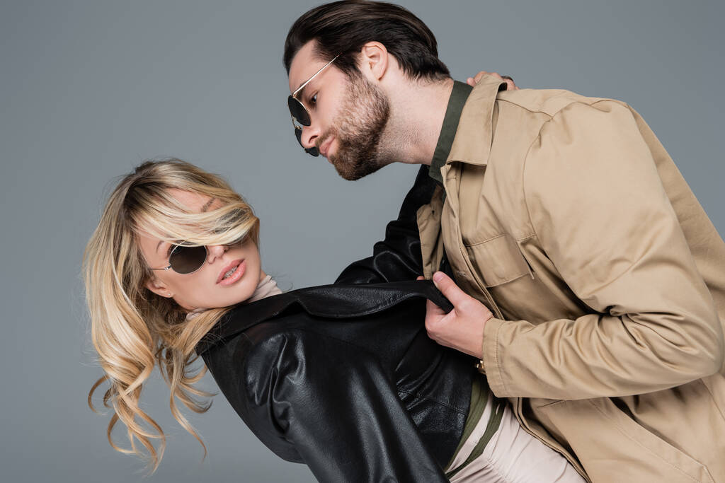 bearded man in sunglasses pulling leather jacket of blonde woman while flirting isolated on grey - Foto, imagen