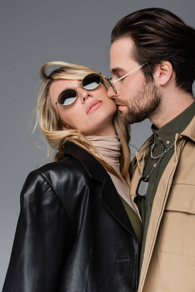 bearded man kissing cheek of blonde and stylish woman in sunglasses isolated on grey - Foto, afbeelding