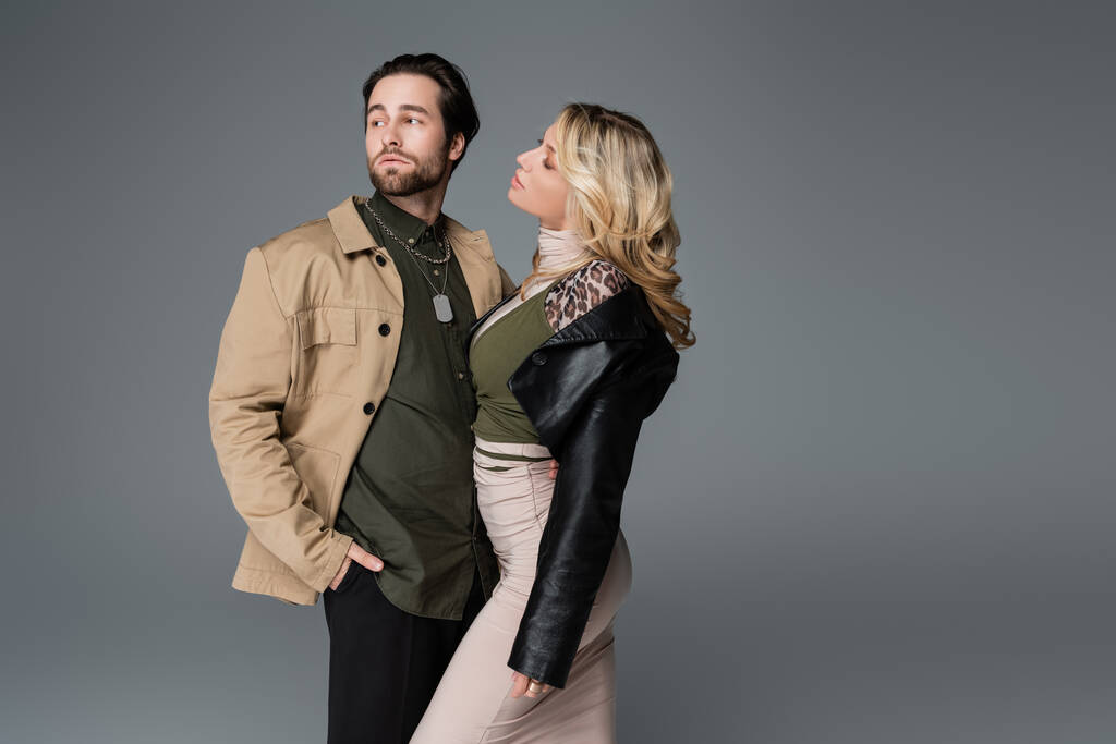 blonde woman in black leather jacket looking at man posing with hand in pocket isolated on grey - Foto, Imagem