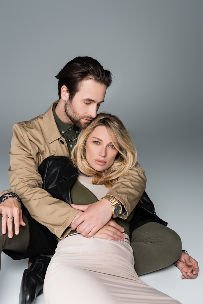 stylish bearded man hugging blonde woman in trendy outfit sitting and looking at camera on grey - Foto, imagen