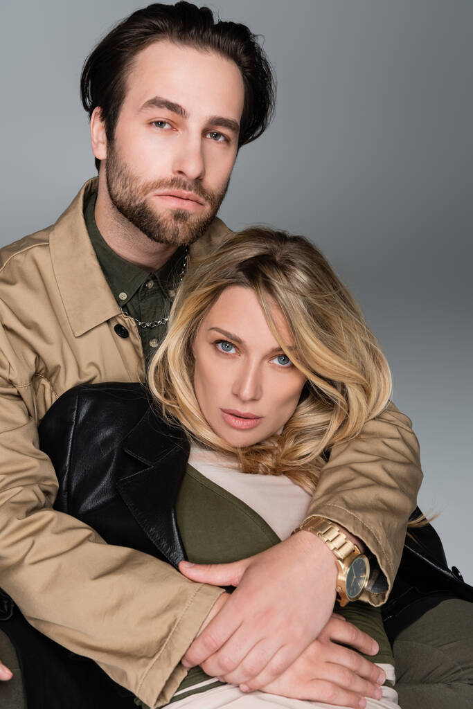  bearded man hugging blonde woman in trendy outfit and looking at camera on grey - Foto, afbeelding