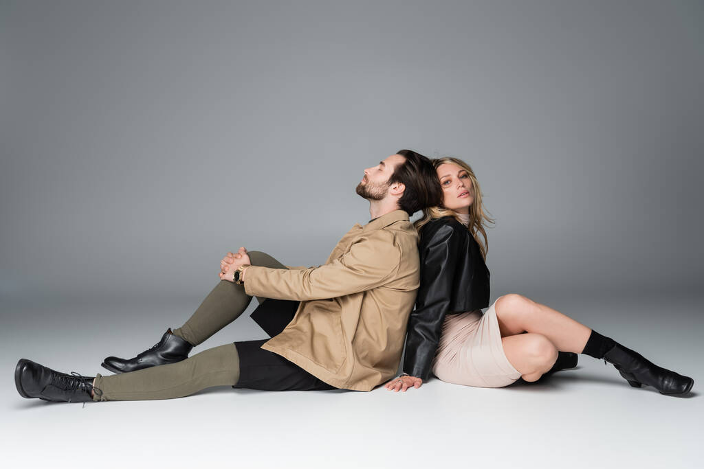 full length of stylish bearded man and blonde woman in trendy outfits sitting back to back on grey - Foto, afbeelding