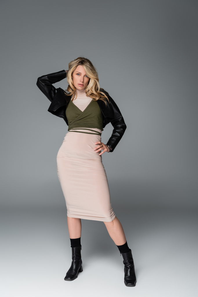 full length of blonde woman in beige dress and black leather jacket posing with hand on hip on grey - Foto, Imagem