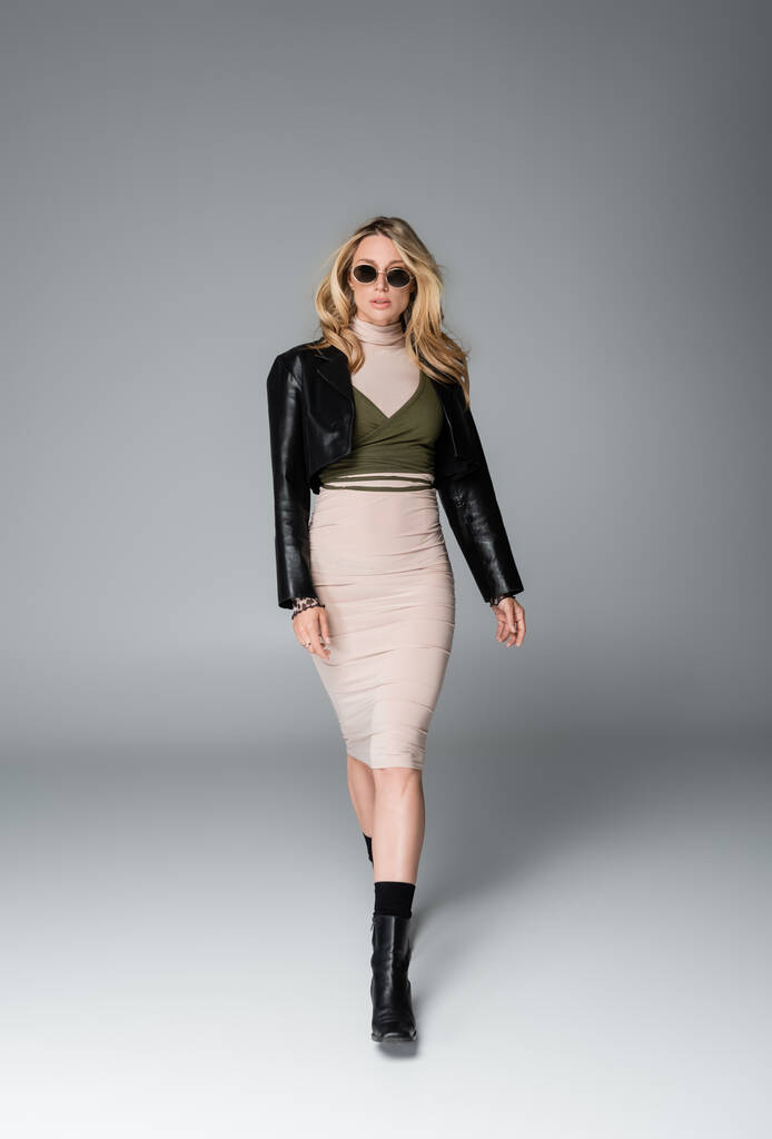 full length of blonde woman in black leather jacket and stylish sunglasses walking on grey - 写真・画像