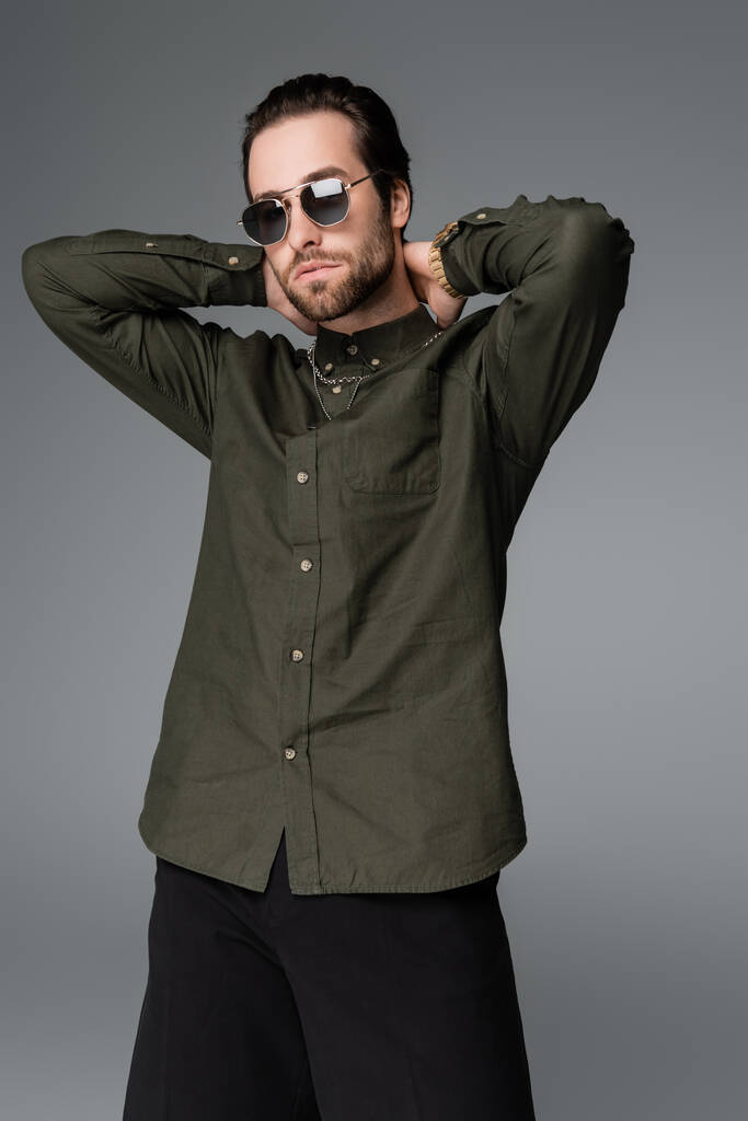γενειοφόρος άνδρας σε κομψό πράσινο πουκάμισο και γυαλιά ηλίου κοιτάζοντας κάμερα, ενώ θέτουν απομονωμένη σε γκρι - Φωτογραφία, εικόνα