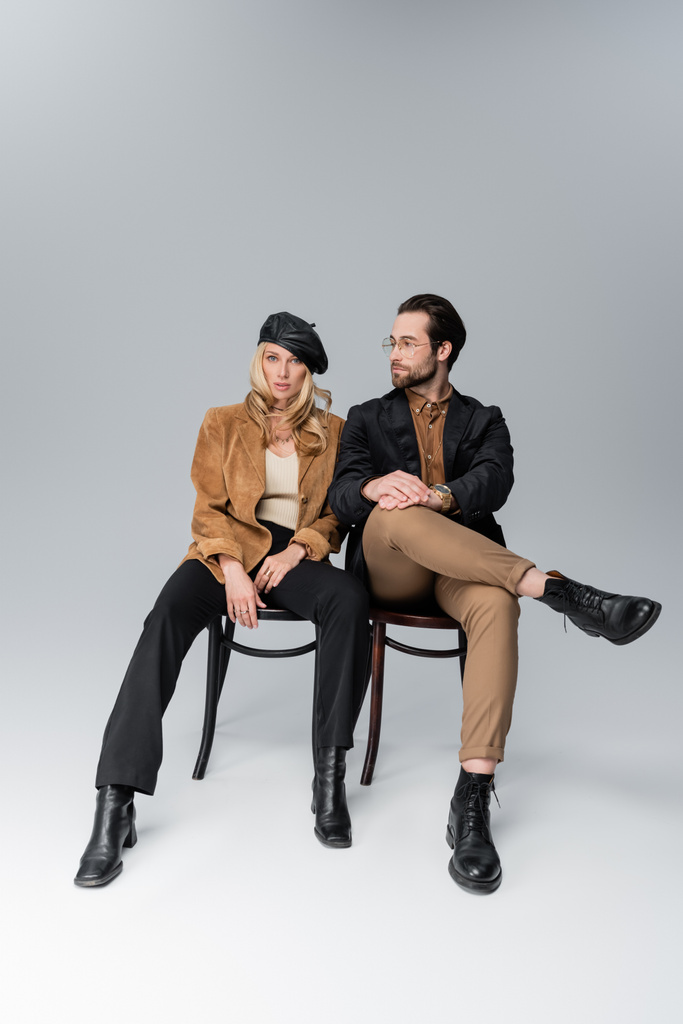full length of blonde woman in stylish beret near bearded man in sunglasses sitting on chairs on grey - Photo, Image