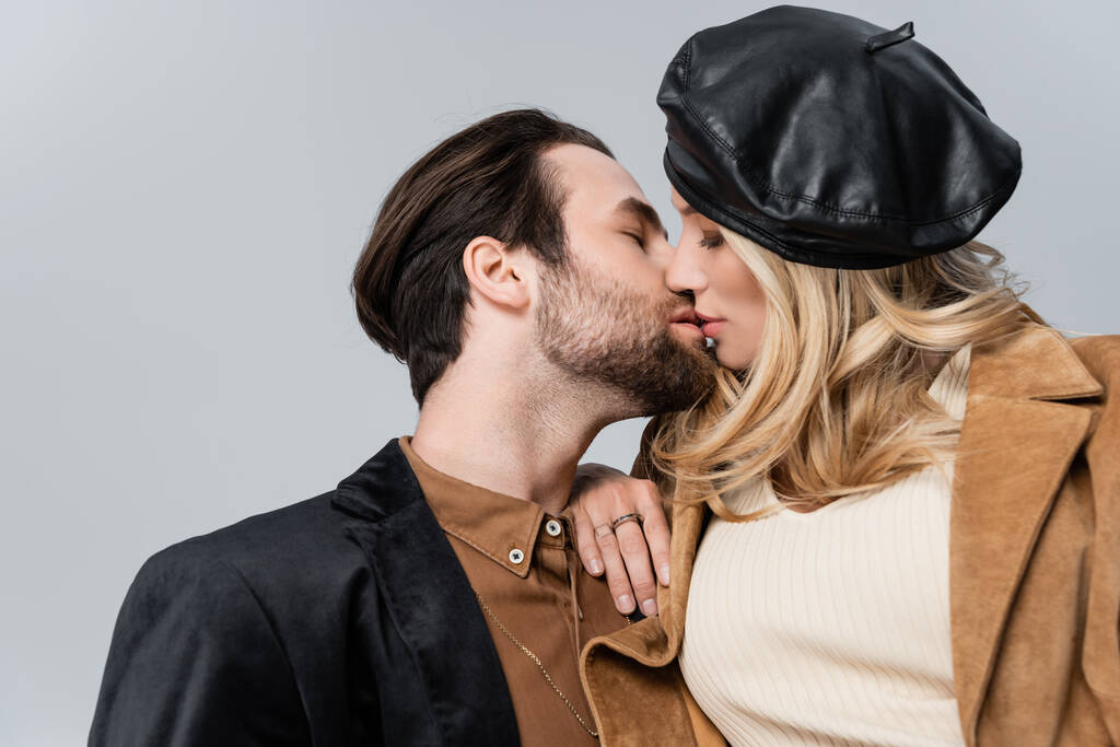 blonde woman in stylish beret kissing bearded man isolated on grey - Foto, imagen