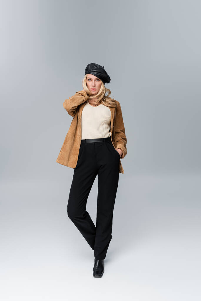 blonde woman in stylish leather beret and beige blazer posing with hand in pocket on grey - 写真・画像
